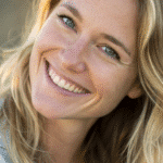 Smiling blonde woman with blue eyes, wearing a casual jacket, looking directly at the camera in natural light.